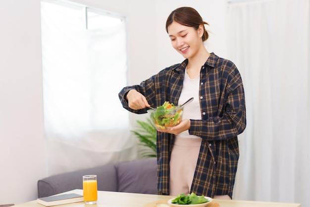 거실 컨셉의 라이프스타일 젊은 아시아 여성이 그릇에 야채 샐러드를 섞기 위해 서 있습니다.