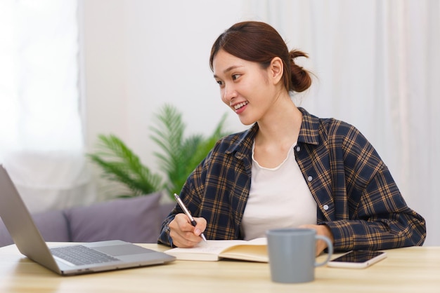 Lifestyle in living room concept Young Asian woman using laptop and taking notes data on notebook