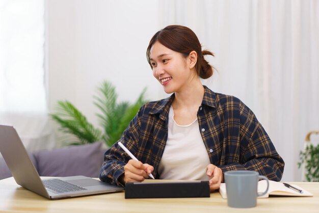 거실 개념의 라이프스타일 젊은 아시아 여성은 노트북을 사용하고 태블릿으로 데이터를 기록합니다.