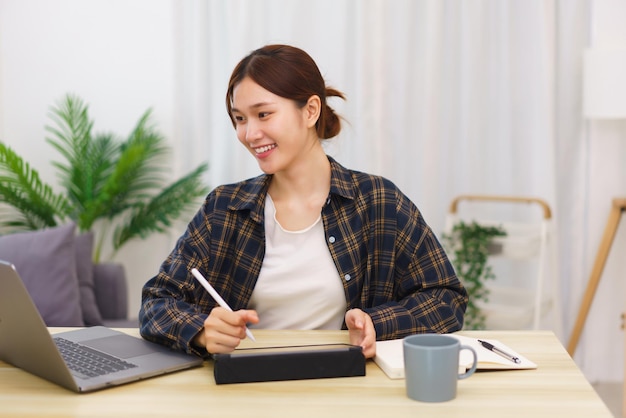 Lifestyle in living room concept Young Asian woman using laptop and taking notes data on tablet