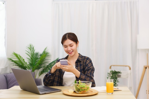 リビングルームのコンセプトのライフスタイルスマートフォンを使用して野菜サラダを食べる若いアジアの女性