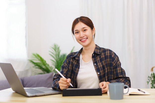 거실 개념의 라이프스타일 젊은 아시아 여성이 집에서 온라인으로 작업하기 위해 태블릿에 글을 쓰고 있습니다.