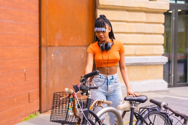 Lifestyle met een jonge trapdanseres in de stad. Zwart meisje van Afrikaanse etnische groep met oranje t-shirt en cowboybroek. De fiets in de stad parkeren bike