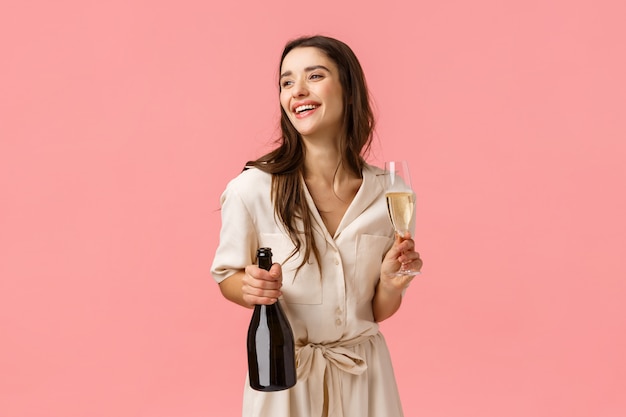 Lifestyle, modern women and beauty concept. Alluring happy young brunette girl in pretty dress, laughing looking aside, open bottle champagne holding glass, drinking and partying