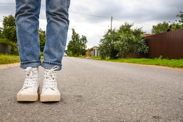 ライフスタイル。ファッショナブルな白いスニーカーでスタイリッシュなブルージーンズのモダンな若い女性が道に立っています。スタイリッシュな婦人靴。若者スタイル。トレンディな靴で女性の足のクローズアップ。