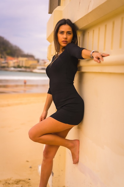 Lifestyle, mooie jonge blanke brunette met een korte zwarte jurk, zomervakantie, camera kijken met sexy look en genieten van het strand