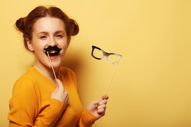 Lifestyle and people concept Happy girl wearing fake mustaches and glasses over yellow background
