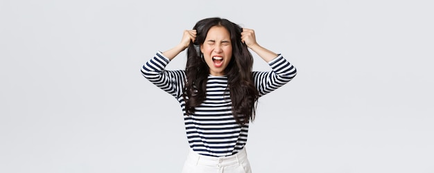 Lifestyle people emotions and casual concept Pissedoff mad and angry asian young woman tossing hair pulling it from head with screams and closed eyes standing bothered white background