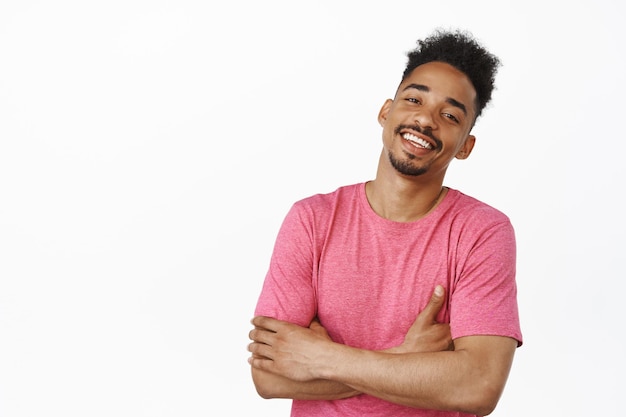 Lifestyle and people. Handsome african american man cross hands on chest, laughing and smiling with carefree expression, standing relaxed in pink casual t-shirt, white background.