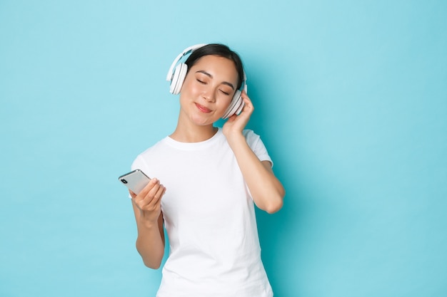 Lifestyle, people and leisure concept. Dreamy beautiful asian girl close eyes and smiling, enjoying listening to music in wireless headphones, dancing to melody, holding smartphone, blue wall