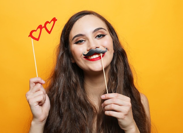 Lifestyle people and party concept Young pretty woman with fake mustaches and glasses over yellow background