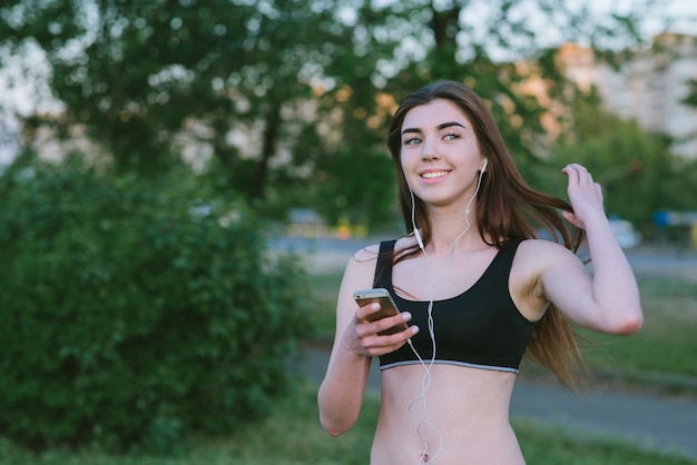 Lifestyle photo of a young smiling girl with a beautiful figure athletic outdoors. Positive athlete with the phone in hand to jog.