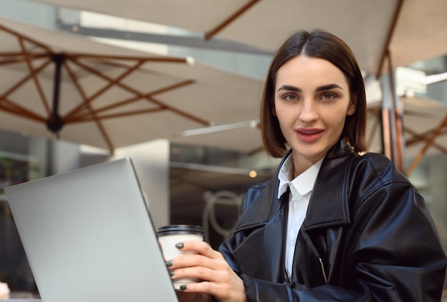 Lifestyle portrait of an attractive brunette woman, copywriter, freelancer with paper cup of takeaway coffee, smiling, looking at camera, working on laptop, sitting at table on summer terrace of cafe.