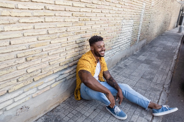 Lifestyle portrait of an attractive and young man with beard smiling and sitting on the floor against a brick wall