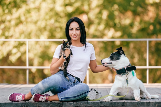 Lifestyle portrait of beautiful young brunette girl with little cat and big hound dog sitting outdoor in park. Happy cheerful smiling teen hugging lovely pets.