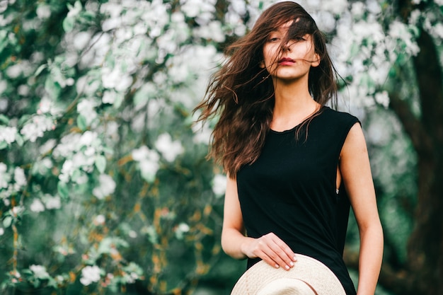 Lifestyle portrait of brunette girl in black dress with straw hat in her hands over blooming apple tree on background.