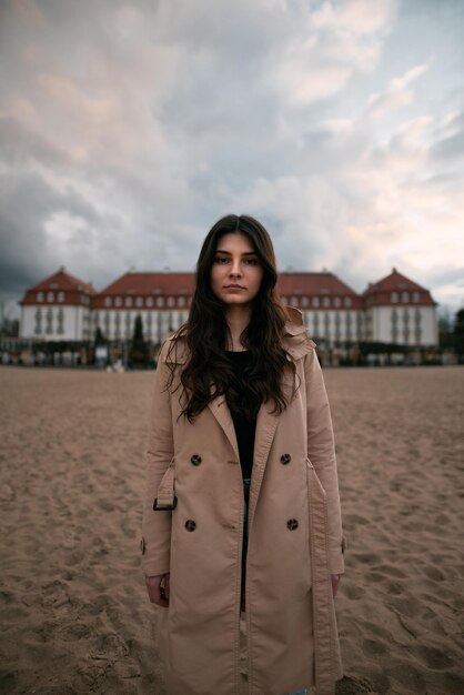 Lifestyle portrait of brunette wears trench coat black shirt and jeans on the ocean coast Girl posing near classical hotel by the seaside during cold time of the year