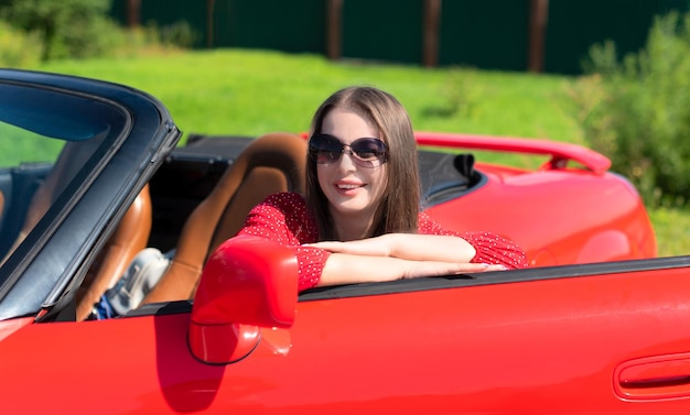 Lifestyle portrait of a carefree beautiful woman in red dress and sunglasses sitting on red cabriolet car and smiling Road trip enjoying freedom concept