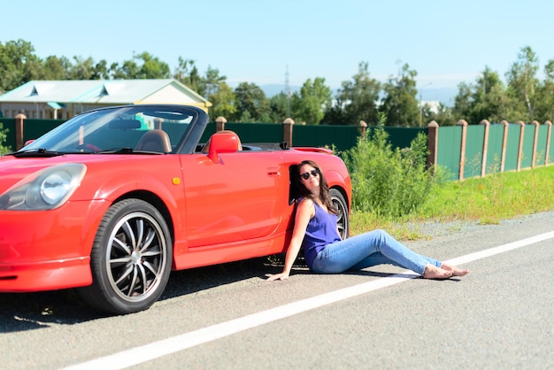 Lifestyle portrait of a carefree beautiful woman in sunglasses dressed casually sitting near red cabriolet car on the roadside Road trip enjoying freedom concept