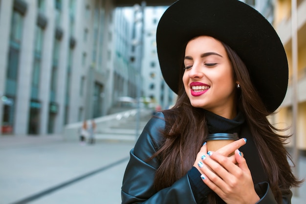 Lifestyle portrait of happy brunette model with cup of coffee walking at the street. Coffee to go concept