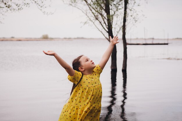 Ritratto di stile di vita di una ragazza gioiosa in un abito giallo con le mani alzate al cielo sul fiume ...