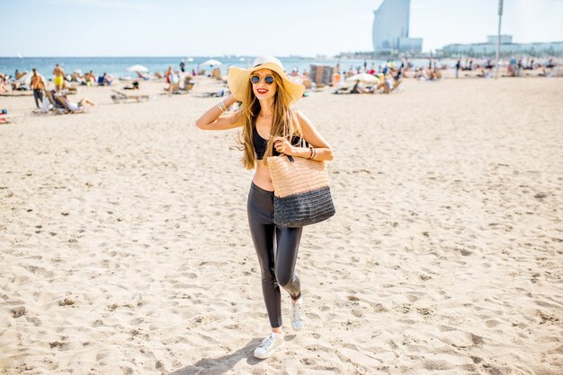 Lifestyle portrait of a pretty woman with sunhat and bag on the beach in Barcelona