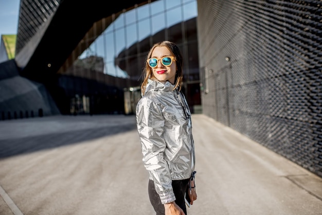 Lifestyle portrait of a stylish woman in silver jacket walking outdoors on the modern architectural background