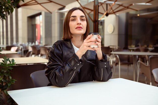 Lifestyle portrait of well-groomed brunette, european woman,\
with cardboard coffee mug looking at camera while resting in summer\
terrace of outside cafeteria restaurant, enjoying autumn cool\
weather