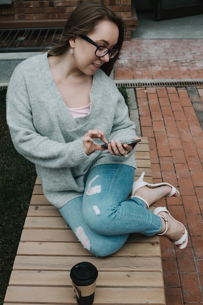 Foto ritratto di stile di vita di giovane smartphone adulto della tenuta della donna bianca con la tazza di caffè da portar via all'aperto, fuoco selettivo