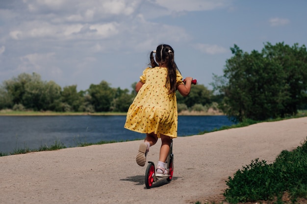 Lifestyle portret achteraanzicht van een meisje in een gele jurk op een scooter op de oever van de rivier in de zomer...