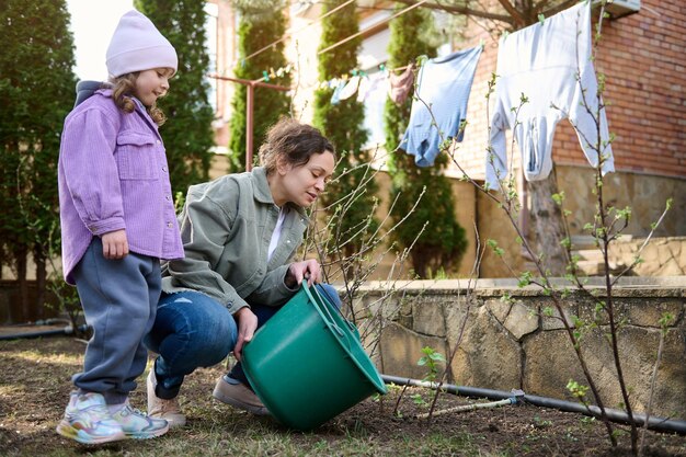 Lifestyle portret nieuwsgierig kind schattig klein meisje kijken naar haar serene moeder jonge struiken en planten water geven in de achtertuin van een landhuis in het voorjaar Tuinbouw tuinieren concept