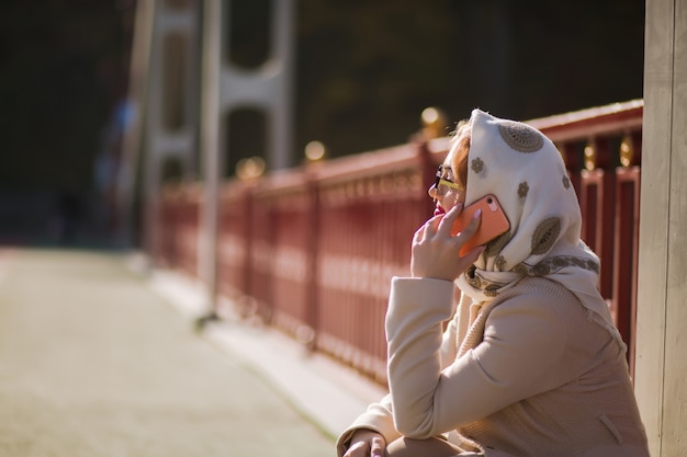 Lifestyle portret van aantrekkelijk blond model draagt beige jas, witte sjaal en praat op haar mobiele telefoon