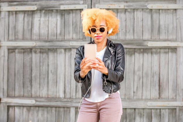 Lifestyle-portret van een Afrikaanse vrouw in leren jas die een selfie-foto maakt met een telefoon die op de achtergrond van de houten muur staat