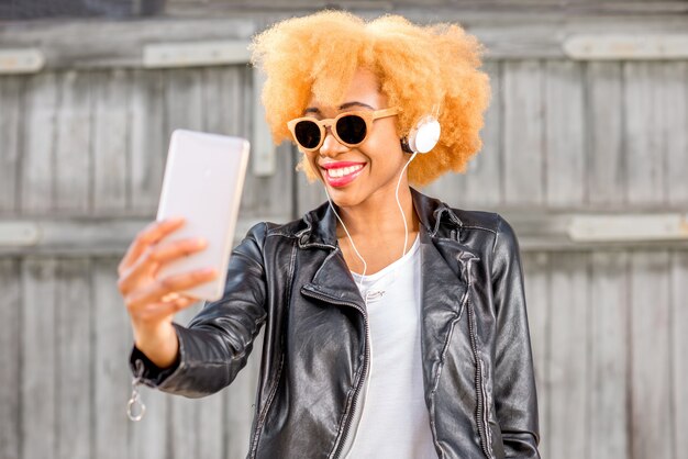 Lifestyle-portret van een Afrikaanse vrouw in leren jas die een selfie-foto maakt met een telefoon die op de achtergrond van de houten muur staat