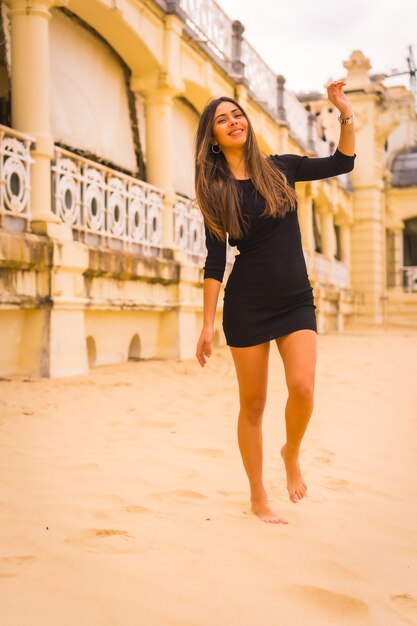 Lifestyle, portret van een jonge brunette blanke vrouw in een korte zwarte jurk, wandelend langs een strand genietend van de zomervakantie