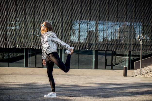 Lifestyle-portret van een stijlvolle vrouw in een zilveren jas die buiten aan het oefenen is in de moderne stedelijke omgeving