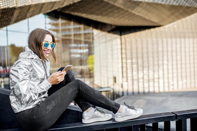 Foto lifestyle-portret van een stijlvolle vrouw in een zilveren jas die buiten met een telefoon zit in de moderne architectonische omgeving