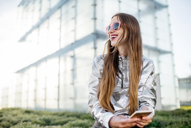 Lifestyle-portret van een stijlvolle vrouw in een zilveren jas die buiten zit in de moderne architectonische omgeving