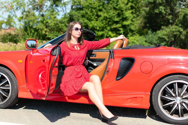 Lifestyle portret van een zorgeloze mooie vrouw in rode jurk en zonnebril zittend op rode cabriolet auto Road trip genieten van vrijheid concept