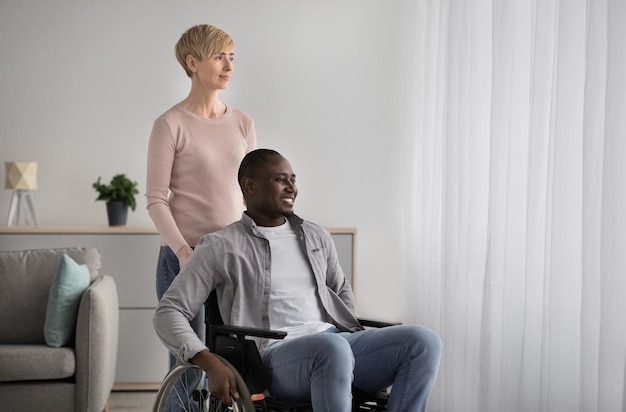 Lifestyle, positief gezin, herstel en ondersteuning van mensen met een beperking thuis en in de kliniek. Vrolijke vrouw van middelbare leeftijd geeft om lachende Afro-Amerikaanse man in rolstoel in woonkamer interieur