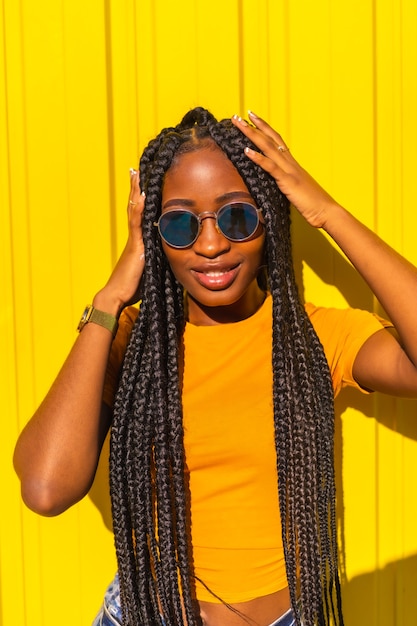라이프 스타일, 긴 머리띠, 노란색 티셔츠 및 노란색 벽에 짧은 청바지가있는 예쁜 흑인 소녀. 선글라스와 함께 웃고