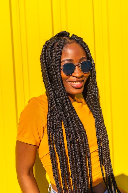 Lifestyle, pretty black girl with long braids, yellow t-shirts and short jeans on a yellow wall. Smiling with sunglasses