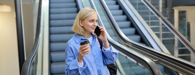 Фото Снимок стиля жизни молодой студентки, разговаривающей по мобильному телефону, пьющей кофе, стоящей рядом