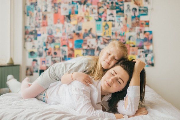 Lifestyle soft focus portrait of happy mom hugs her adorable daughter on white bed. Indoor happy family portrait of smiling mother and her daughter lying together on bed with decorative wall behind.