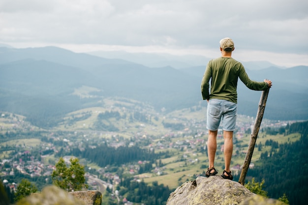 Lifestyle summer portrait from behind of successful man with wooden stick standing on top of mountaing with beautiful landscape in front. 