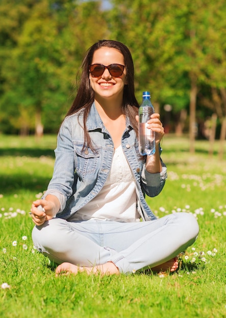생활 방식, 여름, 휴가, 음료, 그리고 사람들의 개념 - 공원 잔디에 물 한 병을 들고 선글라스를 끼고 웃고 있는 어린 소녀