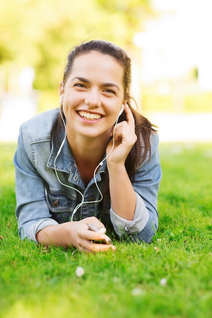 lifestyle, summer vacation, technology, leisure and people concept - smiling young girl with smartphone and earphones lying on grass