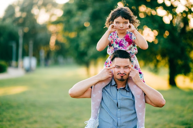 Lifestyle sunset portrait of unusual couple of dad with daughter on shoulders making funny expressive faces. Happy family playing at summer nature outdoor. Fatherhood and childhood. Dad loves his girl