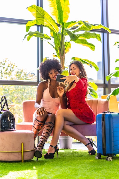 Lifestyle, two friends who have just arrived at the hotel with suitcases taking a selfie with the mobile, a Caucasian blonde girl and a black girl with afro hair, a pink dress and a red dress.