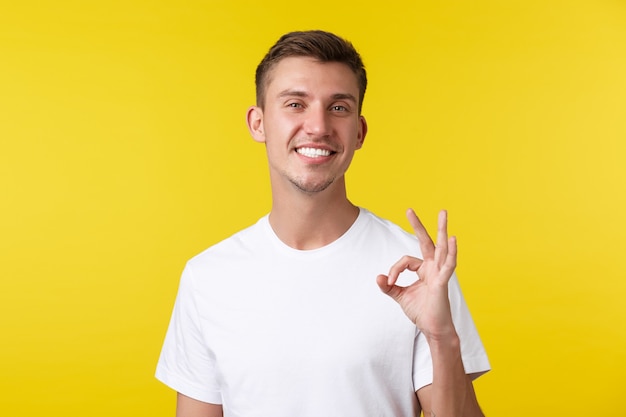 Lifestyle, zomer en mensen emoties concept. Portret van een uitgaande knappe blonde man die tevreden glimlacht, een goed gebaar toont, goedkeurt en van geweldig werk houdt, mooie baan, gele achtergrond.